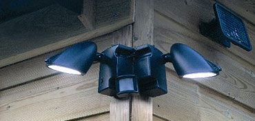 A dual-head outdoor security light is mounted on the corner of a wooden building in NC, illuminating both sides. A small solar panel is visible above the light—demonstrating Pre-eminent Electrical Service Alamance County quality.