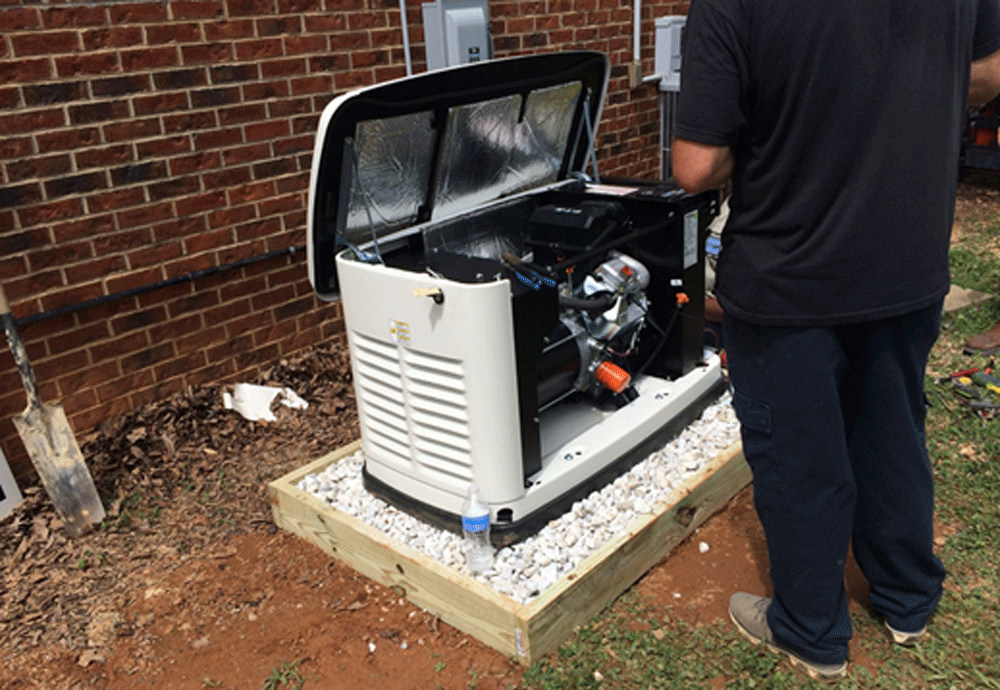 A person stands next to a house, working on an outdoor generator with its cover open, revealing internal components&mdash;a scene showcasing Pre-eminent Electrical Service in Alamance County, NC. The generator sits on a gravel and wooden base near a brick wall.