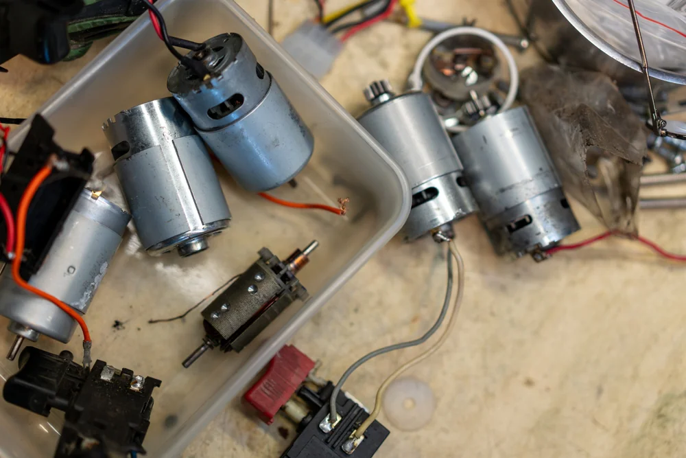 A close-up view of several small electric motors and assorted electronic components scattered on a work surface and inside a plastic container. Wires and connectors are visible among the parts.