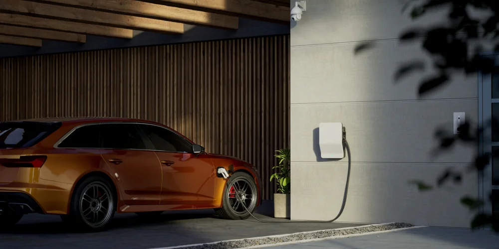 An orange electric car is parked in a modern carport, plugged into a wall-mounted charging station on a light-colored wall, with plants and wooden paneling in the background.