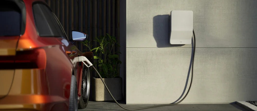 A red electric car is parked beside a white wall-mounted charging station, with the charging cable plugged into the car. A potted plant sits nearby on the ground.