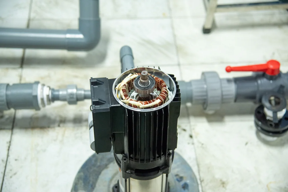 Top view of an electric water pump motor with its cover removed, showing exposed copper windings and internal parts, surrounded by PVC plumbing pipes and a red-handled valve on a tiled floor.