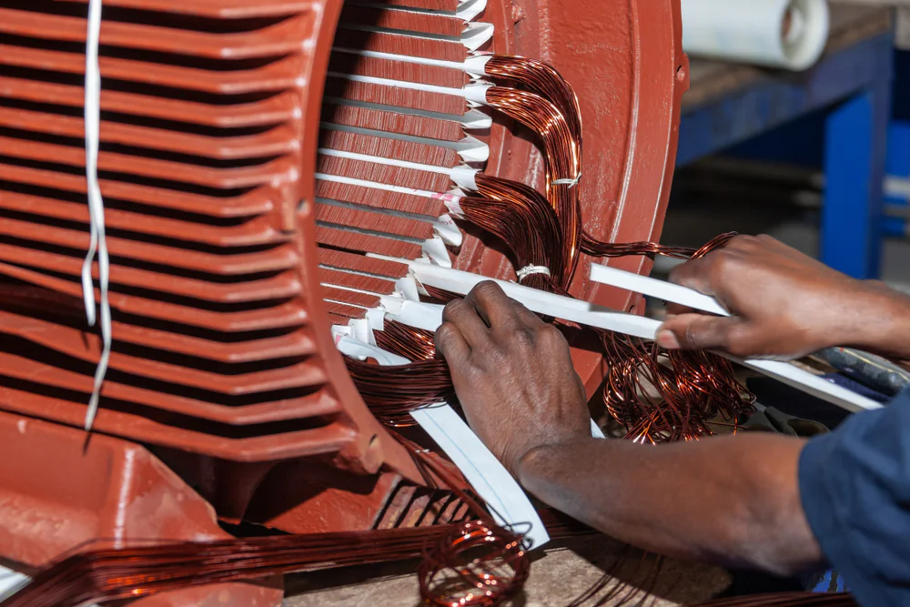 A person’s hands winding copper wires inside the stator of a large electric motor during assembly or repair in an industrial setting.