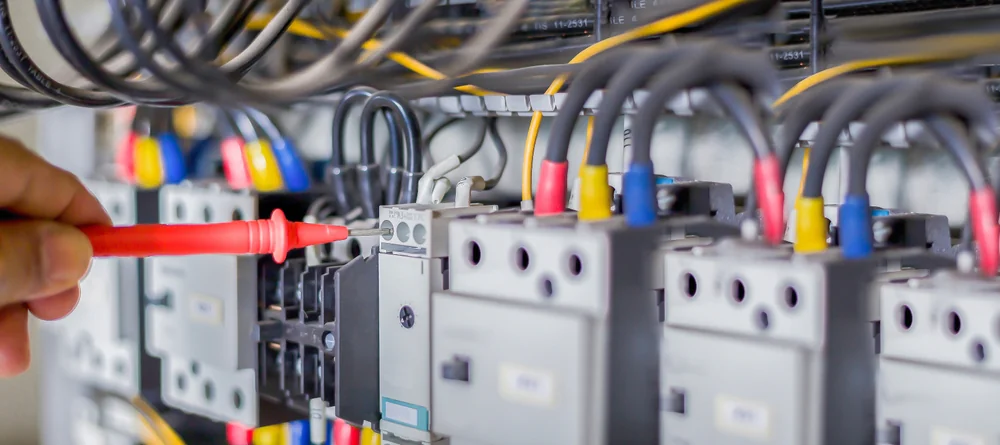 A hand uses a red screwdriver to adjust or repair a component inside an electrical control panel with various wires and connectors visible.