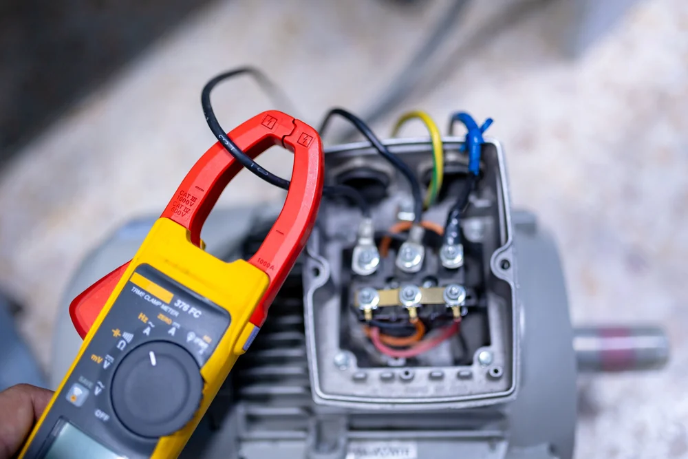 A person holds a digital clamp meter near exposed wires and terminals inside an open electrical motor housing, checking electrical connections.