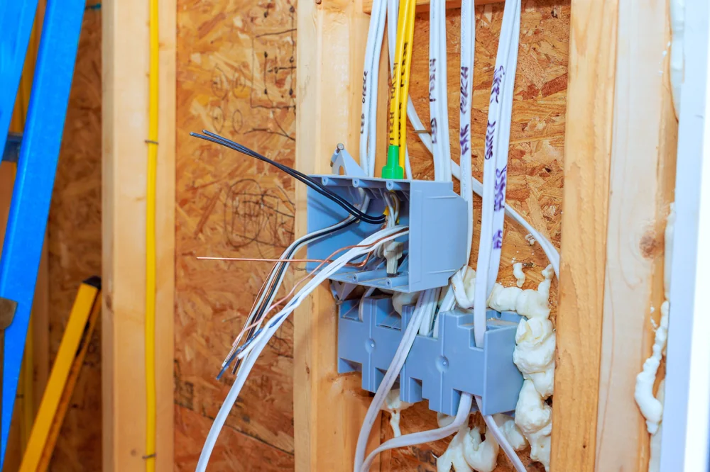 Exposed electrical wiring and junction boxes installed in a wooden wall frame at a construction site, with some wires sticking out and spray foam insulation visible around the boxes.