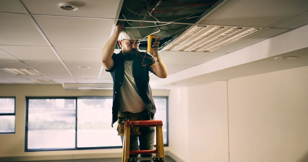 A worker in a hard hat stands on a ladder, inspecting and testing electrical wires in an open ceiling panel of a well-lit office space.