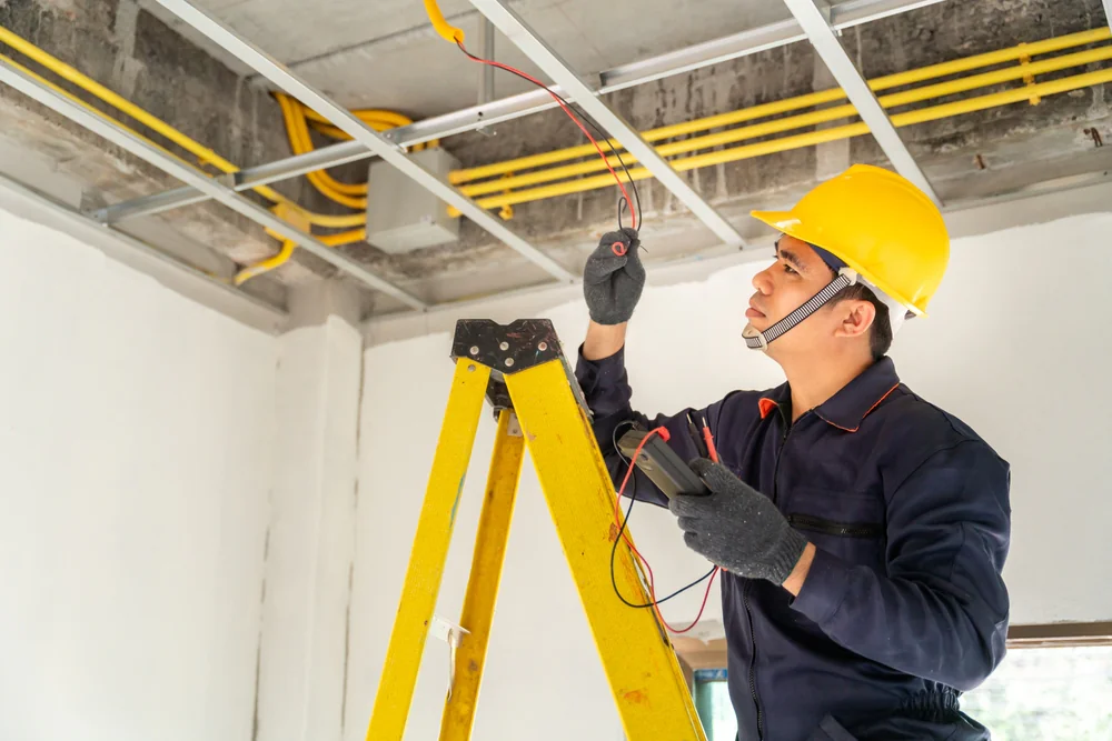 An electrician wearing a yellow hard hat and gloves stands on a ladder, using a multimeter to check electrical wiring in the ceiling of a building under construction.