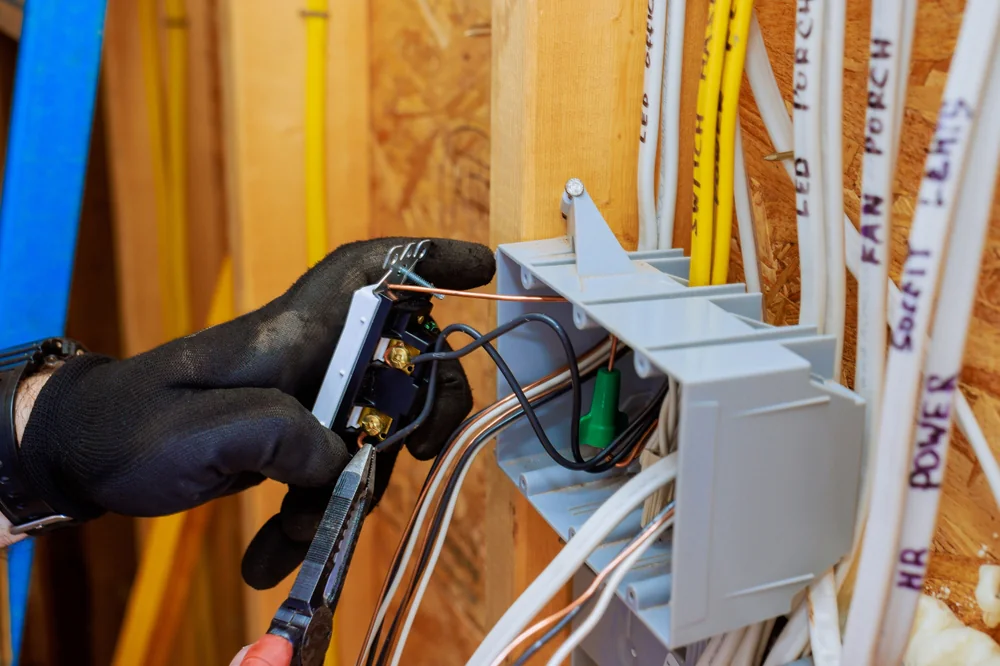 A person wearing black gloves uses pliers to install or repair an electrical outlet, handling wires connected to a junction box in a partially finished wall with visible wiring and insulation.