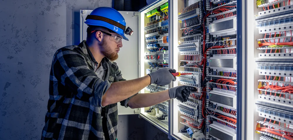 An electrician wearing a blue hard hat, safety glasses, gloves, and a plaid shirt uses a screwdriver to work on complex electrical panels filled with wires inside an industrial setting.