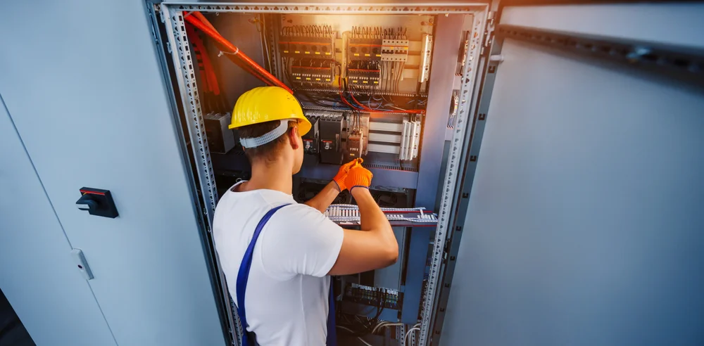 An electrician wearing a yellow hard hat, orange gloves, and a white shirt works on an open electrical control panel, adjusting wiring inside an industrial setting.