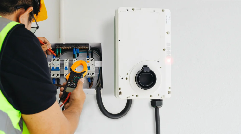 An electrician wearing a safety vest and hard hat checks an electrical panel with a multimeter next to an EV charging station mounted on a white wall.