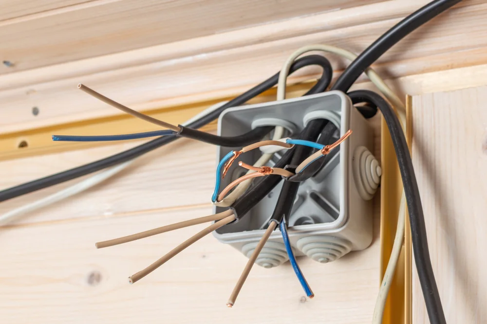 An electrical junction box with multiple exposed wires protruding, including black, blue, and brown wires, mounted on a wooden wall. The wires are not yet connected or covered for safety.