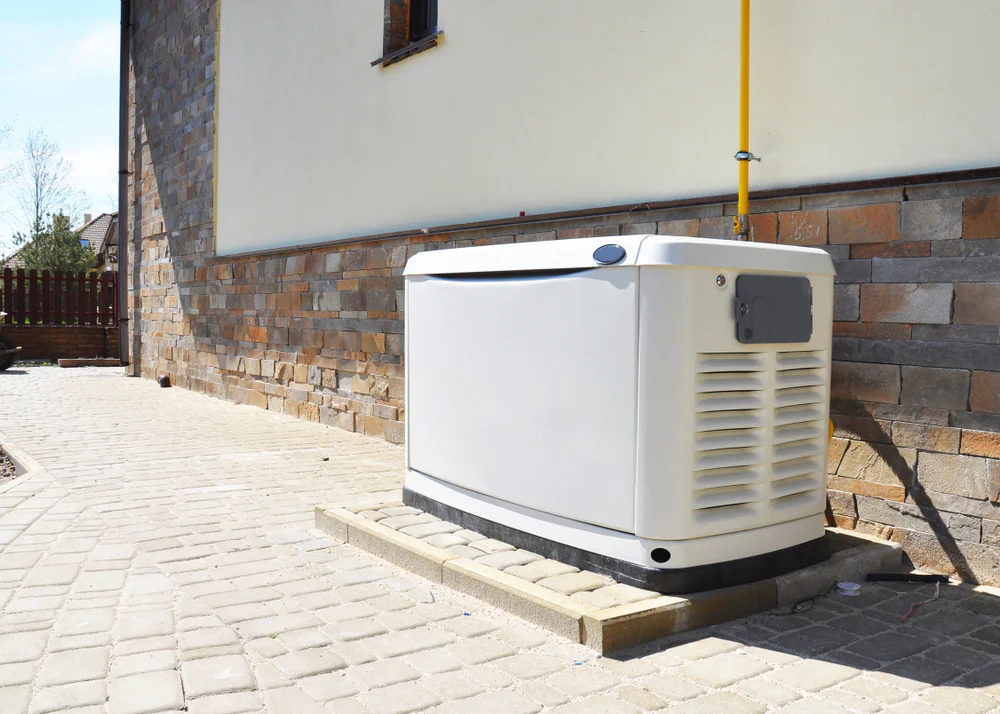 A standby generator sits on a concrete pad next to a house with stone and brick siding on a paved walkway, connected by a yellow gas line.