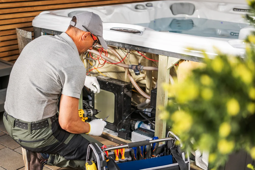 A technician in a gray shirt and cap uses a power drill to repair the wiring beneath an outdoor hot tub, with tools spread out nearby and plants visible in the foreground.