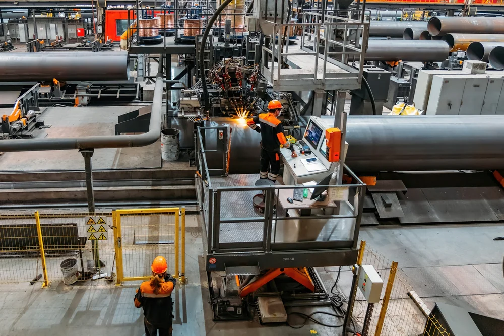 Two workers in safety gear operate machinery in a large industrial factory, with metal pipes and automated equipment visible. One worker is at a control panel; sparks fly from a welding station nearby.