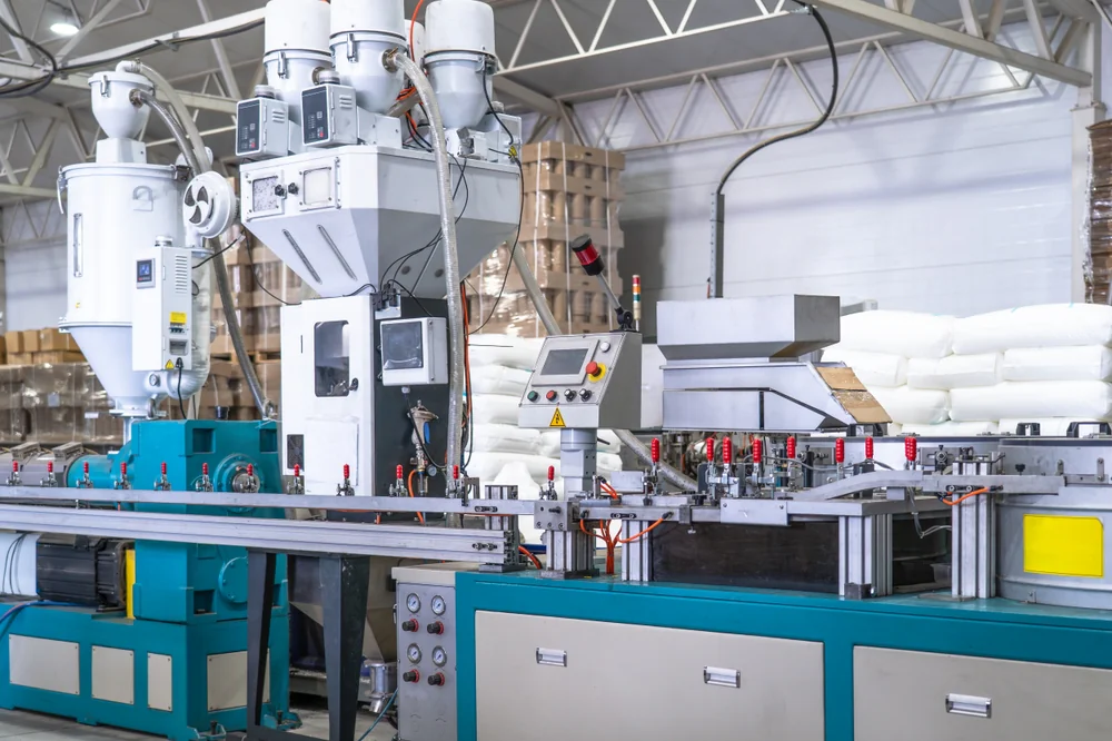 A modern plastic extrusion machine operates in a factory setting, with control panels, pipes, and large bags of raw plastic pellets stacked nearby. The environment is clean and industrial.
