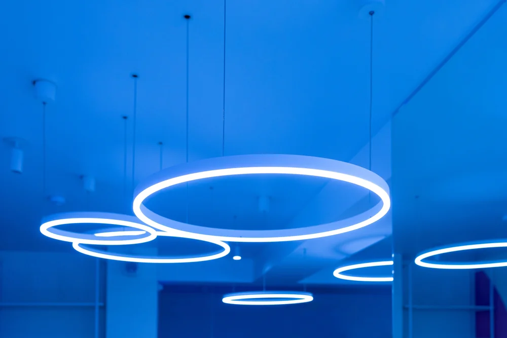 Modern ceiling lights in circular ring shapes glow with a blue hue, suspended from a smooth ceiling by thin wires, creating a futuristic and minimalistic ambiance in the room.
