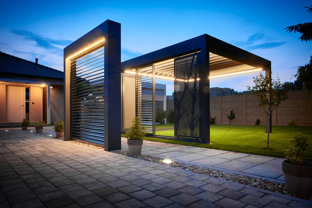 Modern outdoor patio with a sleek pergola featuring integrated lighting and metal slats. The area is surrounded by potted plants, a stone walkway, and a grassy yard, all under a twilight sky.