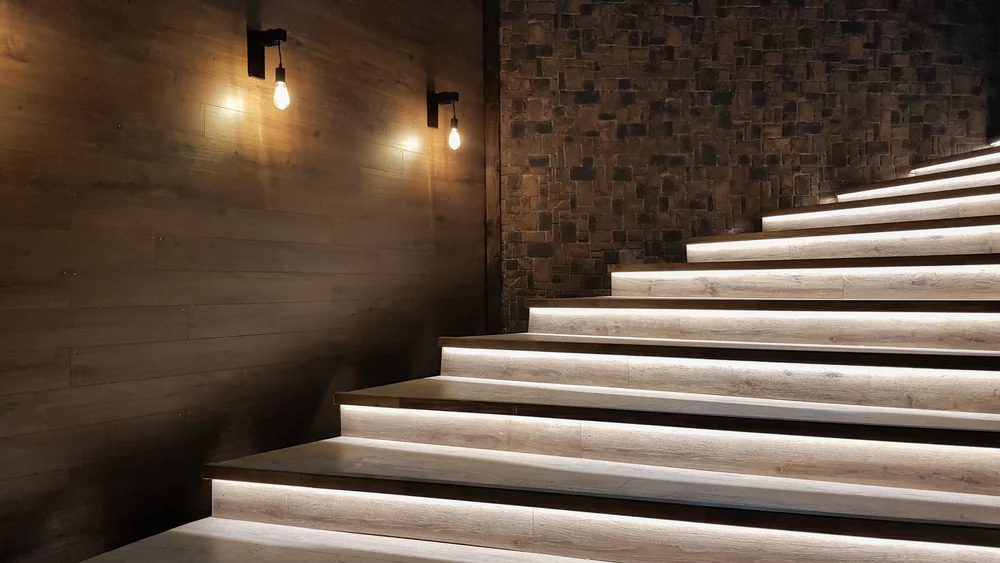 A modern wooden staircase with illuminated steps, set against a textured brick wall. Two exposed light bulbs are mounted on the wooden wall, creating a warm and inviting atmosphere.