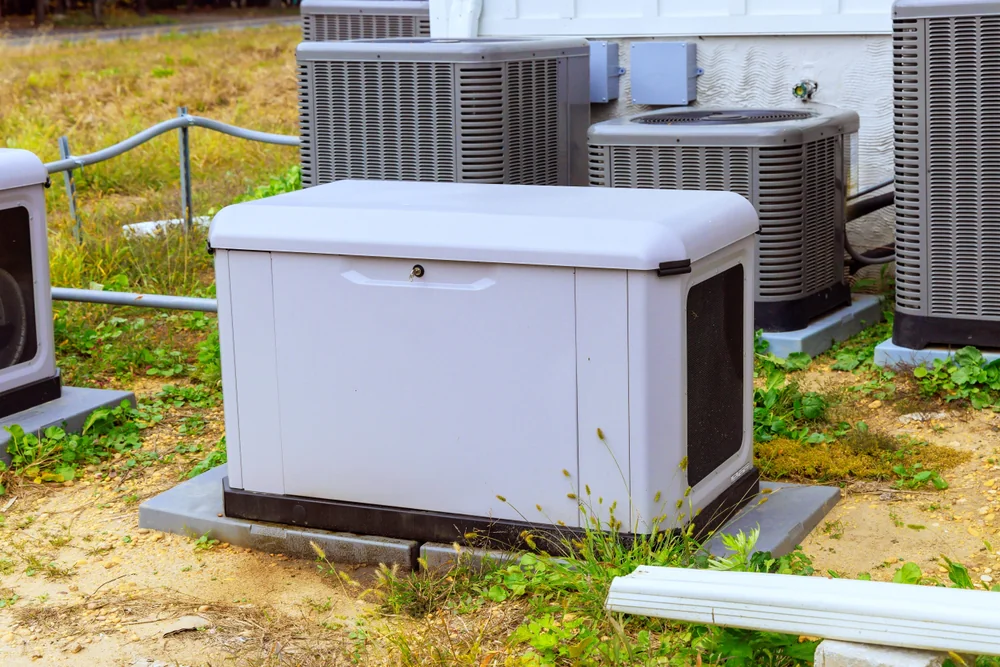 A standby backup generator sits on a concrete pad outdoors, surrounded by three air conditioning units and grass near a building.
