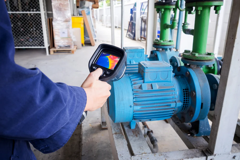 A person uses a handheld thermal imaging camera to inspect a blue industrial electric motor connected to green pipes in a facility. The camera screen displays the motor’s heat signature in various colors.
