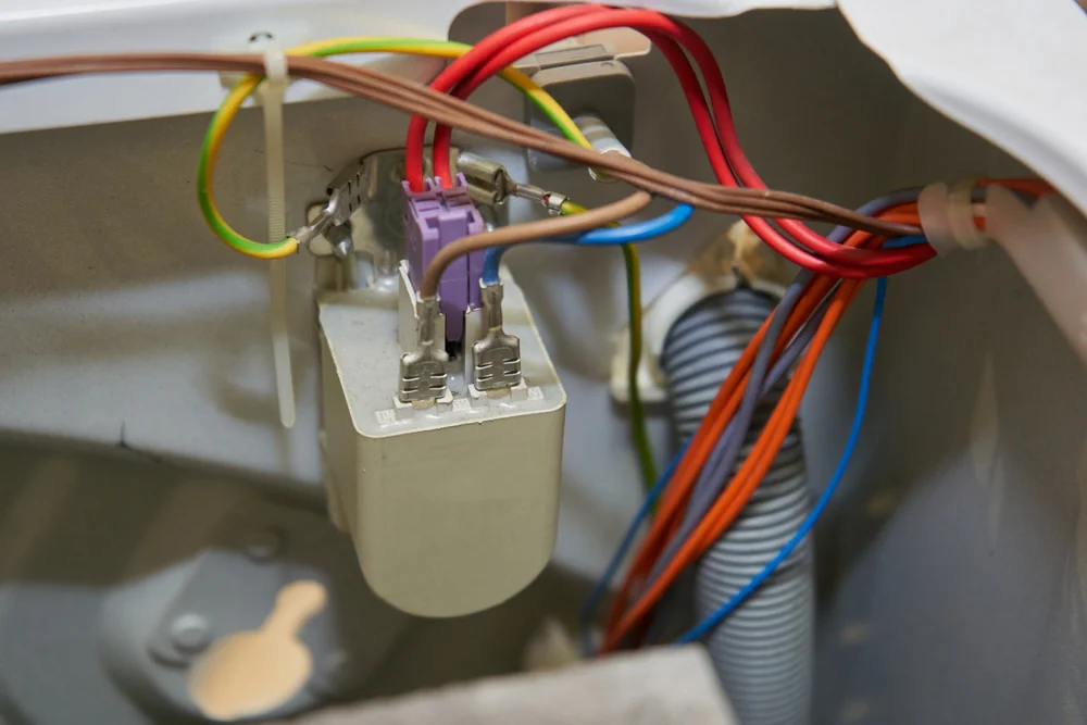 Close-up of electrical wires connected to terminals on a component inside an appliance, possibly a washing machine, with various colored wires and a gray hose visible.