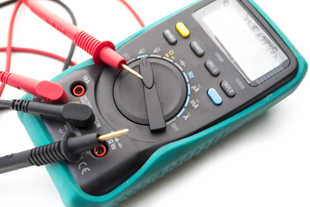 A digital multimeter with red and black probes inserted, resting on a white surface. The device has a green protective cover and a central dial for selecting measurement modes.