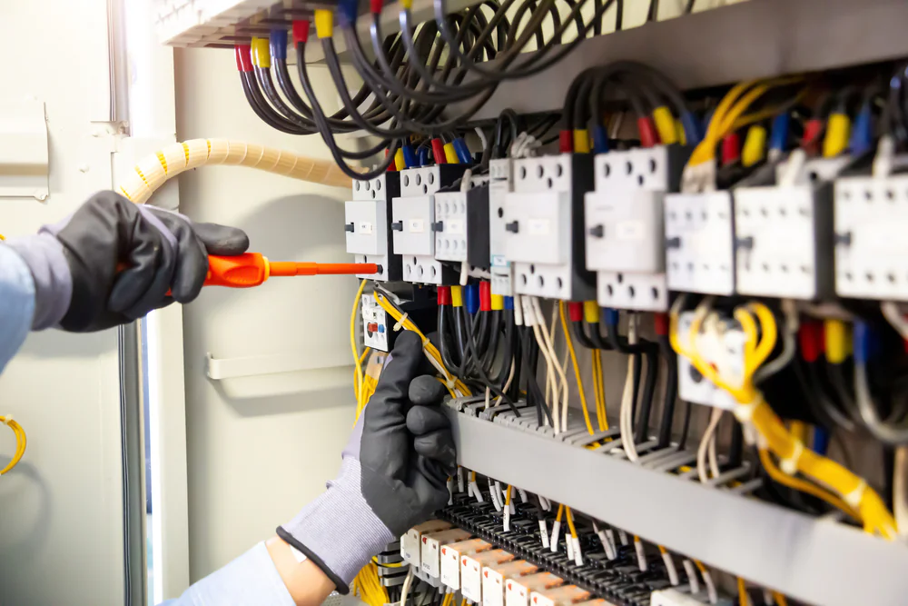 A person wearing gloves uses tools to work on electrical wiring inside a control panel, connecting or adjusting yellow and black wires among various switches and components.