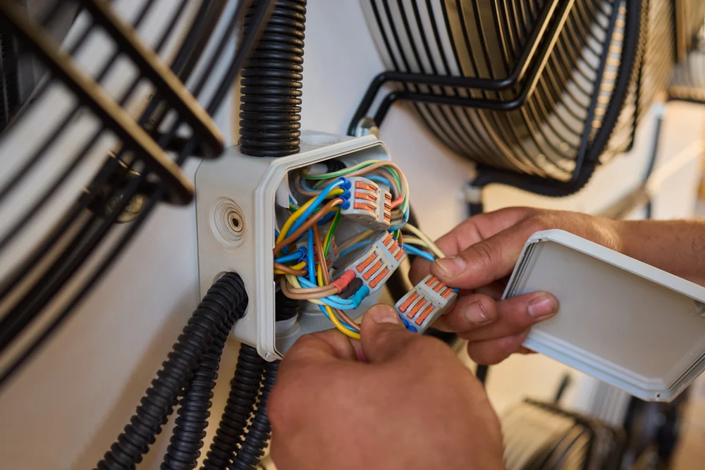 A person&rsquo;s hands connect colored electrical wires inside a white junction box, surrounded by black cable conduits and large metal fan covers.