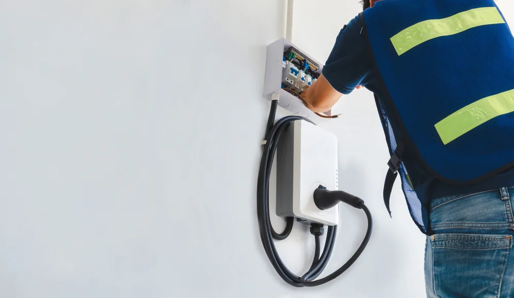 A person wearing a blue safety vest is installing or repairing an electric vehicle charging station mounted on a white wall. The station has a cable and plug attached.