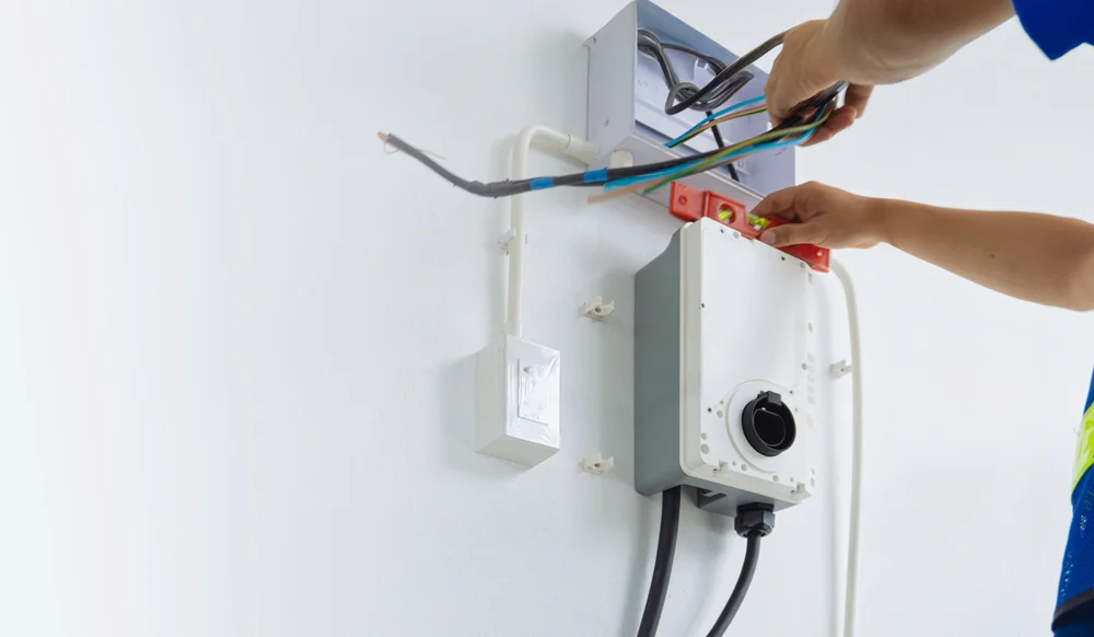 An electrician installs or repairs wiring for a wall-mounted electrical box, using tools and a level, with cables and conduit visible against a white wall.