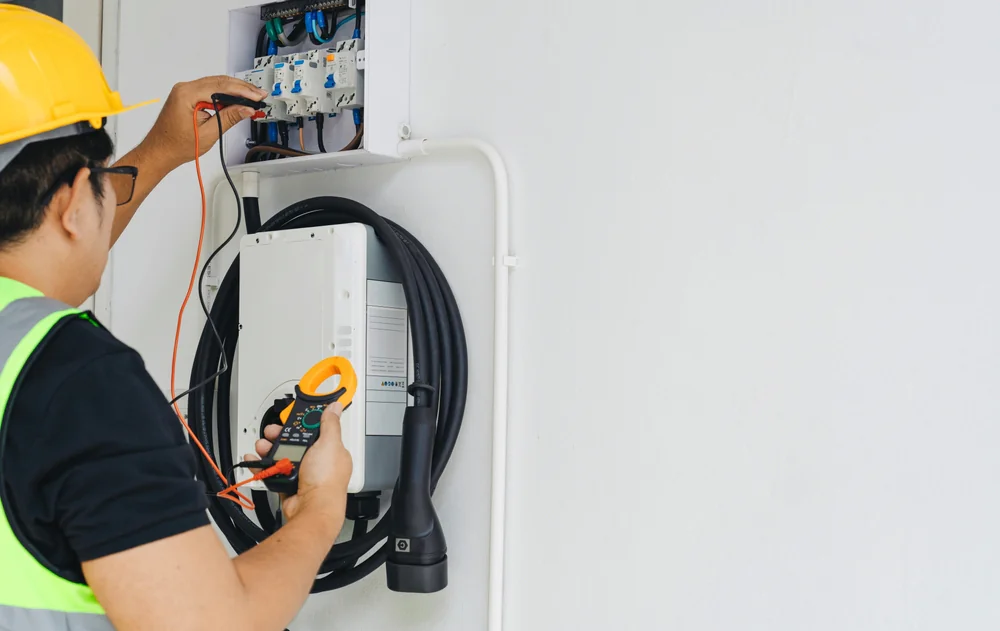 An electrician wearing a yellow hard hat and safety vest tests electrical connections with tools at a wall-mounted control panel, with cables and equipment visible.