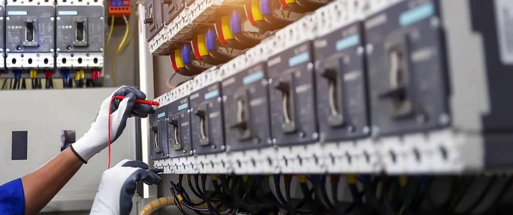 A person wearing white gloves uses a multimeter to check connections inside an electrical control panel filled with switches, wires, and circuit breakers.