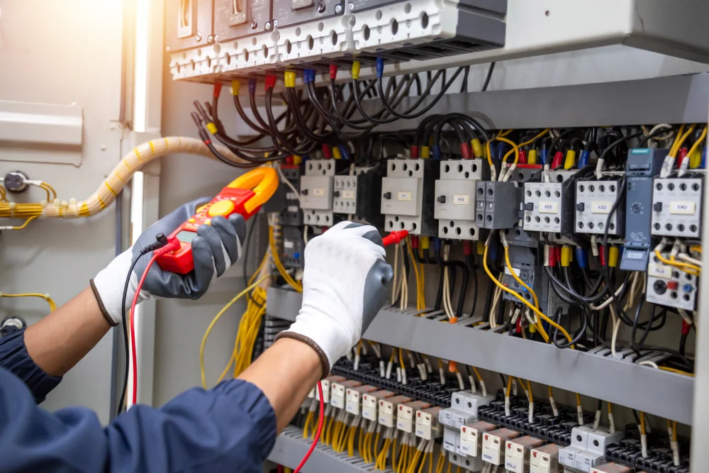 A person wearing white gloves uses a handheld multimeter to check electrical wiring inside an open control panel filled with wires, switches, and circuit breakers.