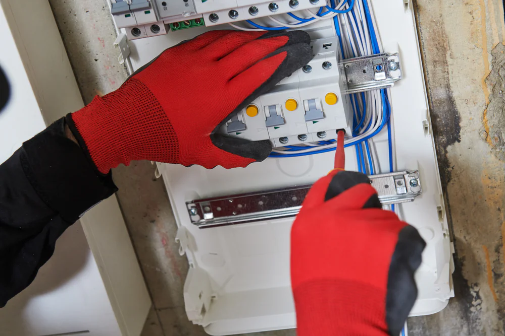 A person wearing red and black gloves uses a screwdriver to work on an electrical panel with blue wires and circuit breakers.
