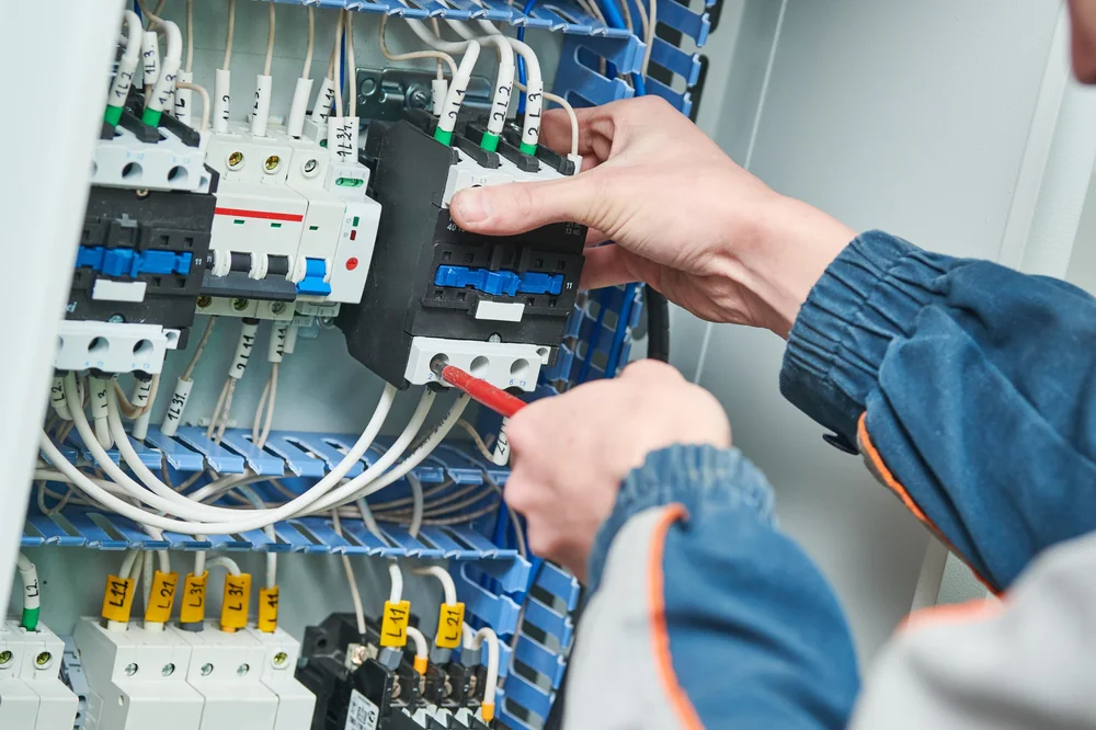 A person in a work uniform uses a screwdriver to adjust or install wiring components inside an electrical control panel filled with switches, wires, and circuit breakers.