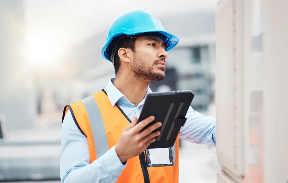 A man wearing a blue hard hat and orange safety vest inspects equipment outdoors while holding a black tablet, suggesting he is conducting a technical or safety inspection on a worksite.