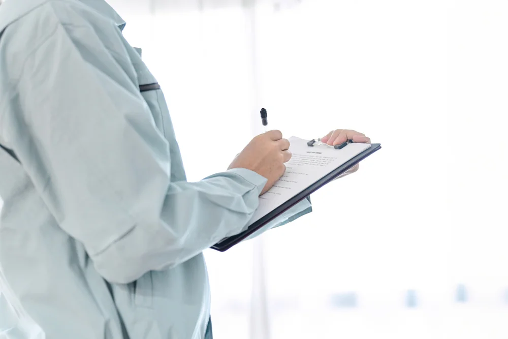 A person in light blue work attire holds a clipboard and writes with a pen, standing in a brightly lit, indoor setting with a white background.