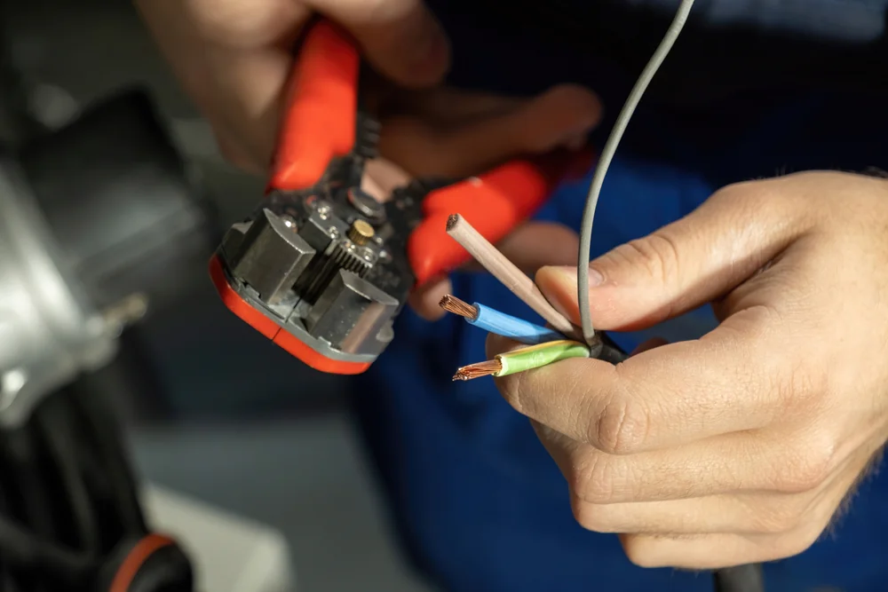 Close-up of hands using red wire strippers to strip insulation from electrical wires, revealing copper conductors inside. The person is holding three wires: blue, green-yellow, and brown.