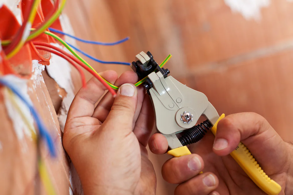 A person uses a wire stripper tool to strip insulation from colored electrical wires inside a wall, preparing them for installation or repair.