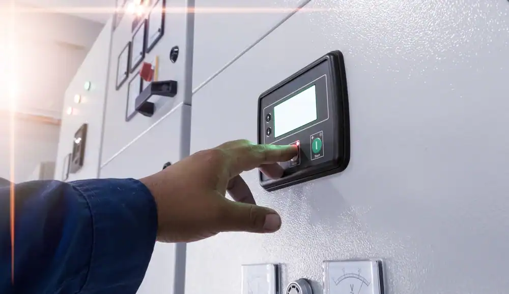 A person wearing a blue sleeve presses a red button on an electronic control panel mounted on a white wall, part of a setup serviced by the pre-eminent electrical service company in Alamance County, NC. Other switches and indicator lights are visible nearby.