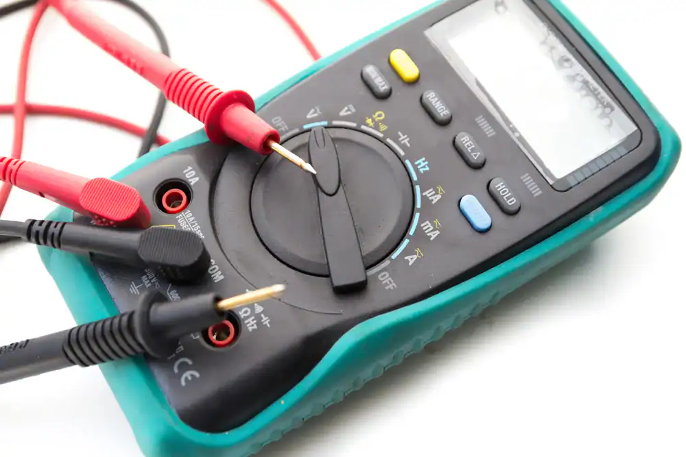 A digital multimeter with a teal protective casing, used by the pre-eminent electrical service company in Alamance County, NC, has its red and black test probes inserted into the ports, displaying various measurement settings on the dial. The display screen is currently off.