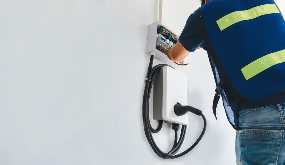 A technician from a pre-eminent electrical service company in Alamance County, NC, wearing a blue safety vest, installs or repairs a wall-mounted electric vehicle charging station with a coiled charging cable hanging from the unit.