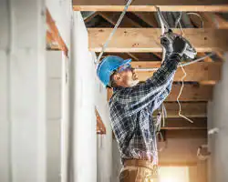 A construction worker wearing a blue hard hat and safety glasses installs electrical wiring on a wooden ceiling frame inside a building under construction for NC’s pre-eminent electrical service company in Alamance County.