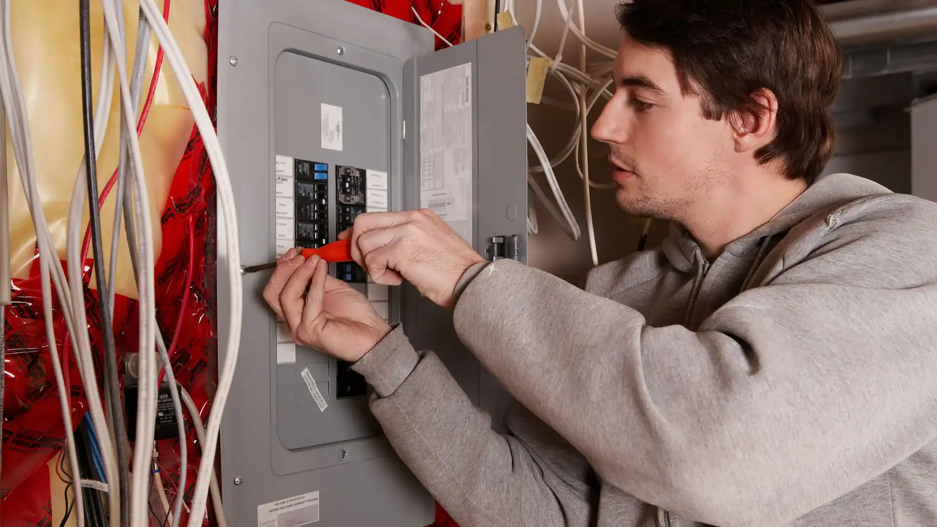 A man in a gray hoodie uses a screwdriver to work on circuit breakers inside an open electrical panel, surrounded by exposed wires and red insulation—demonstrating the expertise of a pre-eminent electrical service company in Alamance County, NC.