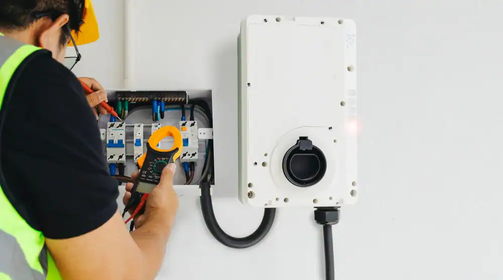 An electrician from a pre-eminent electrical service company in Alamance County, NC, wearing a safety vest and helmet, uses a multimeter to check electrical connections in an open panel by an EV charging station enclosure.