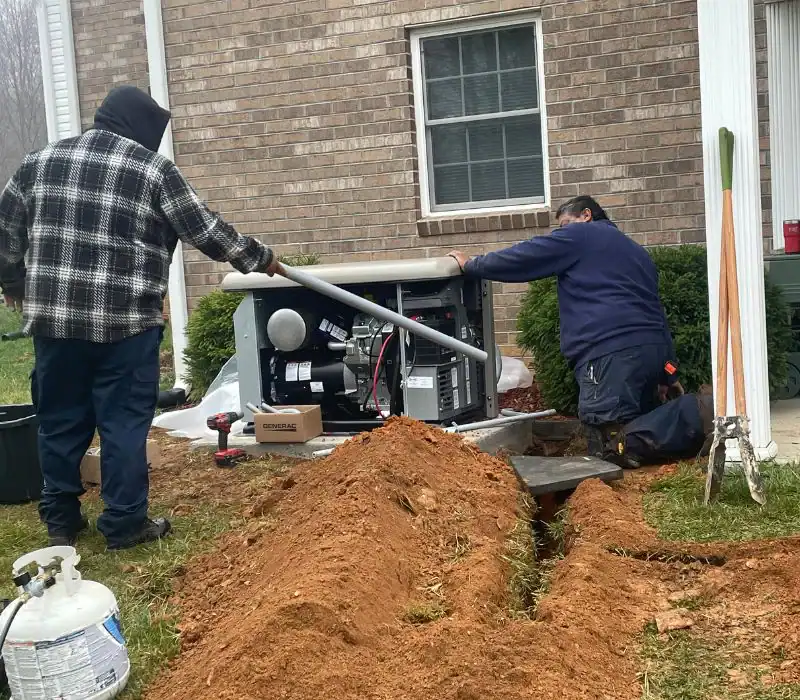 Two people from a pre-eminent electrical service company in Alamance County, NC, install a large generator outside a brick house. Tools, pipes, and a propane tank are nearby. A trench leads from the house to the generator. Shrubs line the wall behind them.