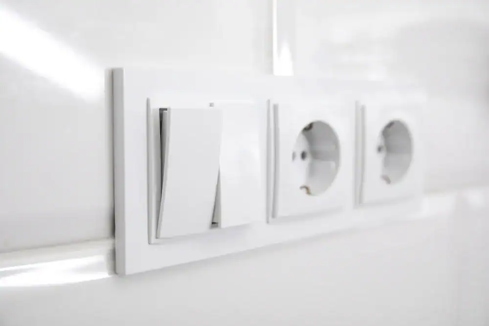 Close-up of a white wall panel with a light switch and two European-style outlets, expertly installed by the pre-eminent electrical service company in Alamance County, NC, mounted on a white tiled wall.