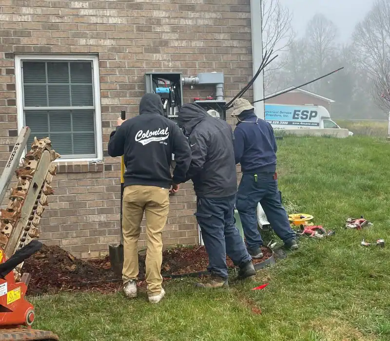 Three people in jackets work by an electrical meter on a brick building, tools and supplies on the grass nearby. One features a "Colonial" logo—this pre-eminent electrical service company serves Alamance County, NC. A foggy business sign is visible.